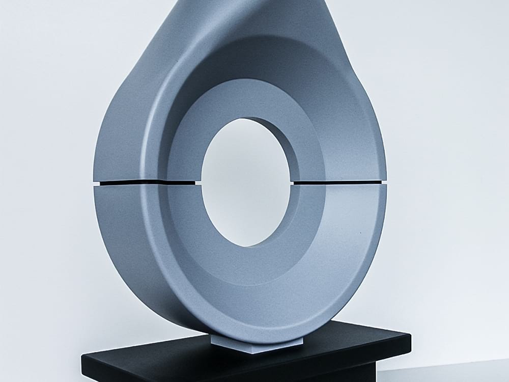 Zweiteiliges Gießereimodell in grauer Farbe mit einer tropfenförmigen Außenkontur und zentraler runder Aussparung. Das Modell steht auf einem schwarzen Präsentationssockel und zeigt eine präzise, symmetrische Formgebung mit glatter Oberfläche. Die Trennfuge zwischen den beiden Hälften ist klar sichtbar und deutet auf eine Formteilverbindung im Gussprozess hin.
