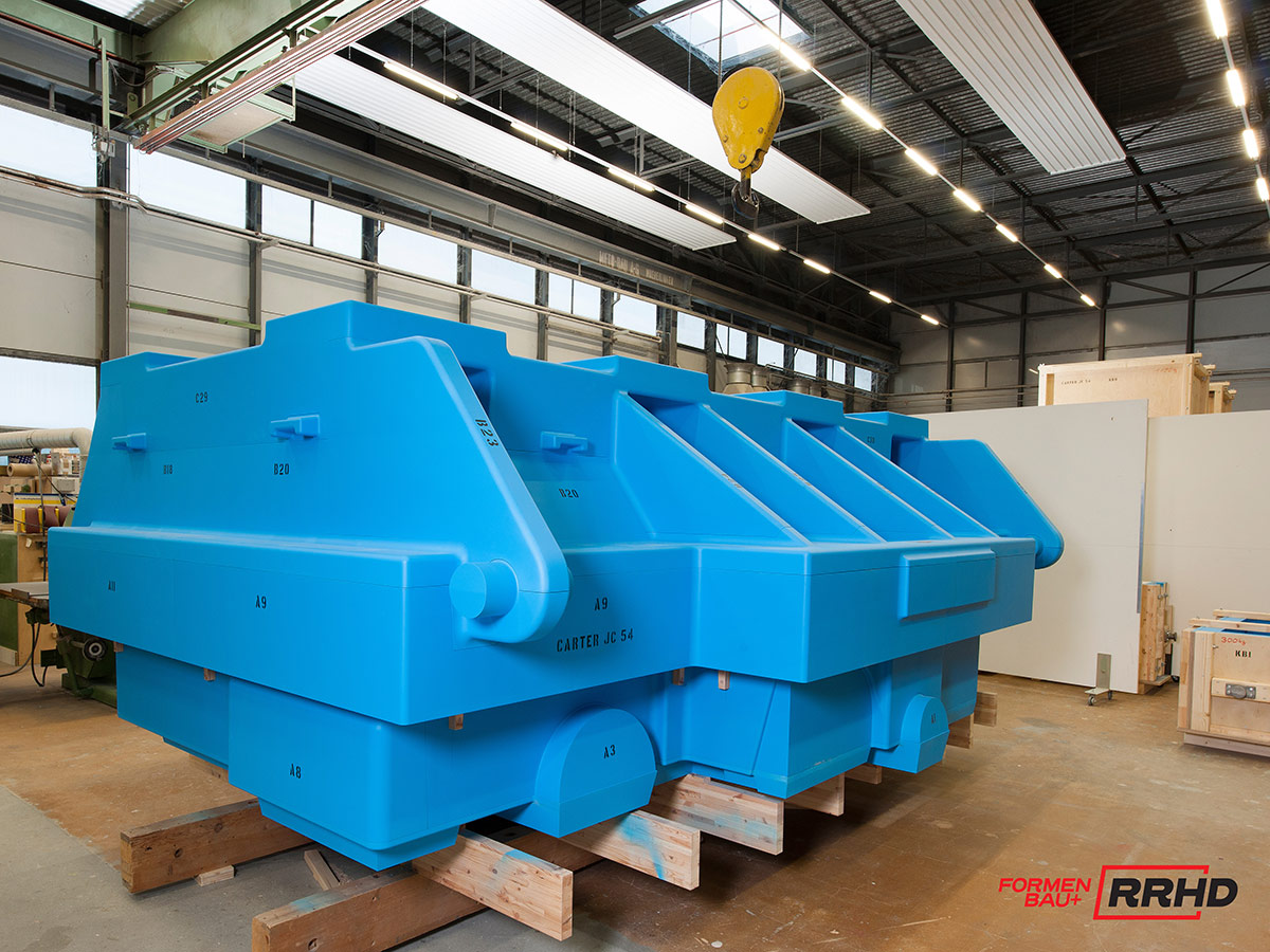 Großes, blaues Gießereimodell in komplexer geometrischer Form, gelagert auf Holzpaletten in einer Werkhalle. Das Modell besteht aus mehreren zusammengefügten Segmenten mit technischen Konturen, Ausrundungen und Verstärkungen. Im Hintergrund sind Hallentore, Maschinen und ein gelber Kranhaken zu sehen.