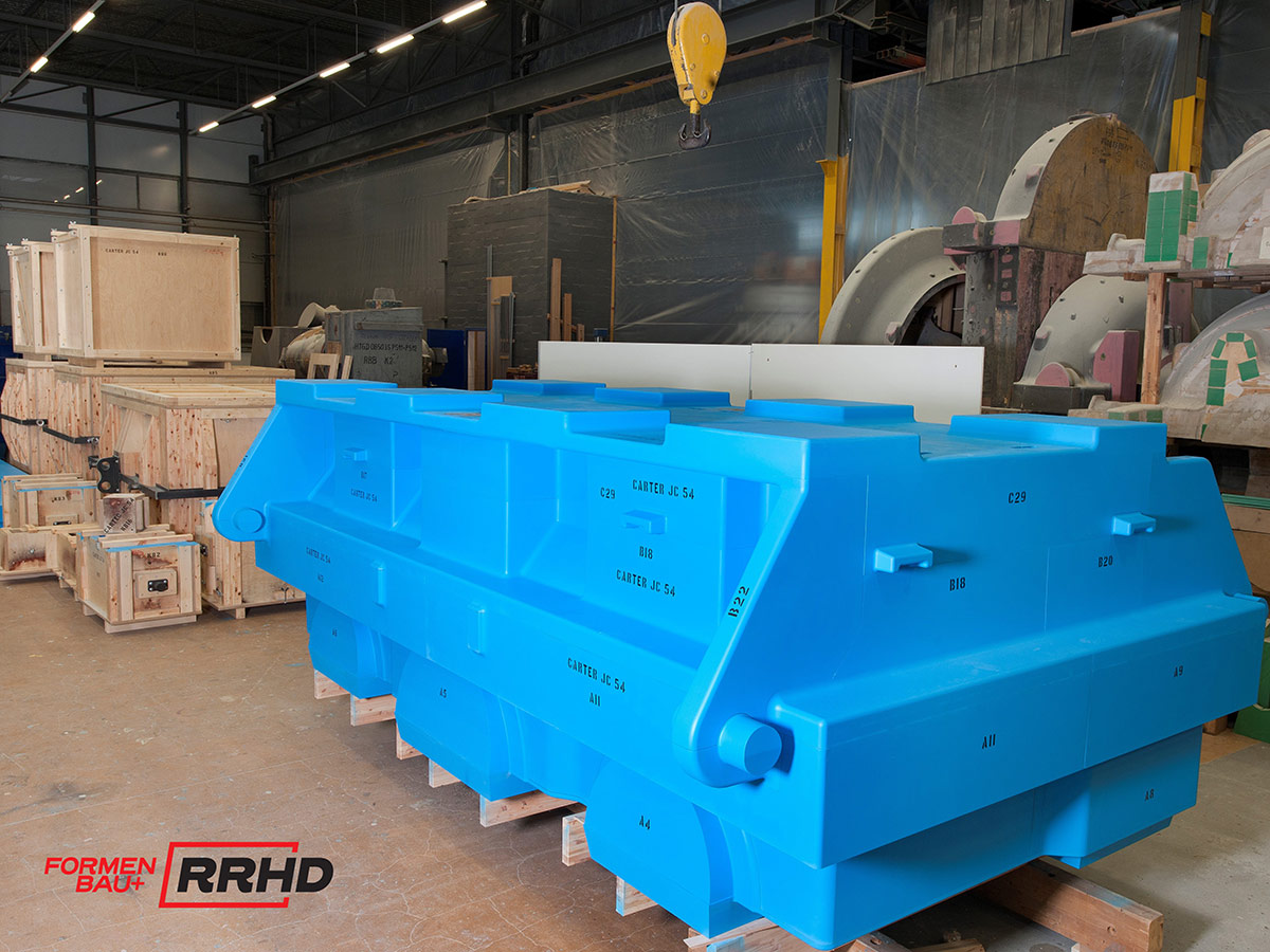 Großes, blau lackiertes Gießereimodell mit komplexer Geometrie und technischen Beschriftungen, gelagert auf Holzunterlagen in einer Werkhalle. Das Modell besteht aus mehreren Segmenten mit klar definierten Flächen, Ausnehmungen und Verstärkungen. Im Hintergrund stehen Kernkästen, weitere Modelle und ein gelber Hallenkran.