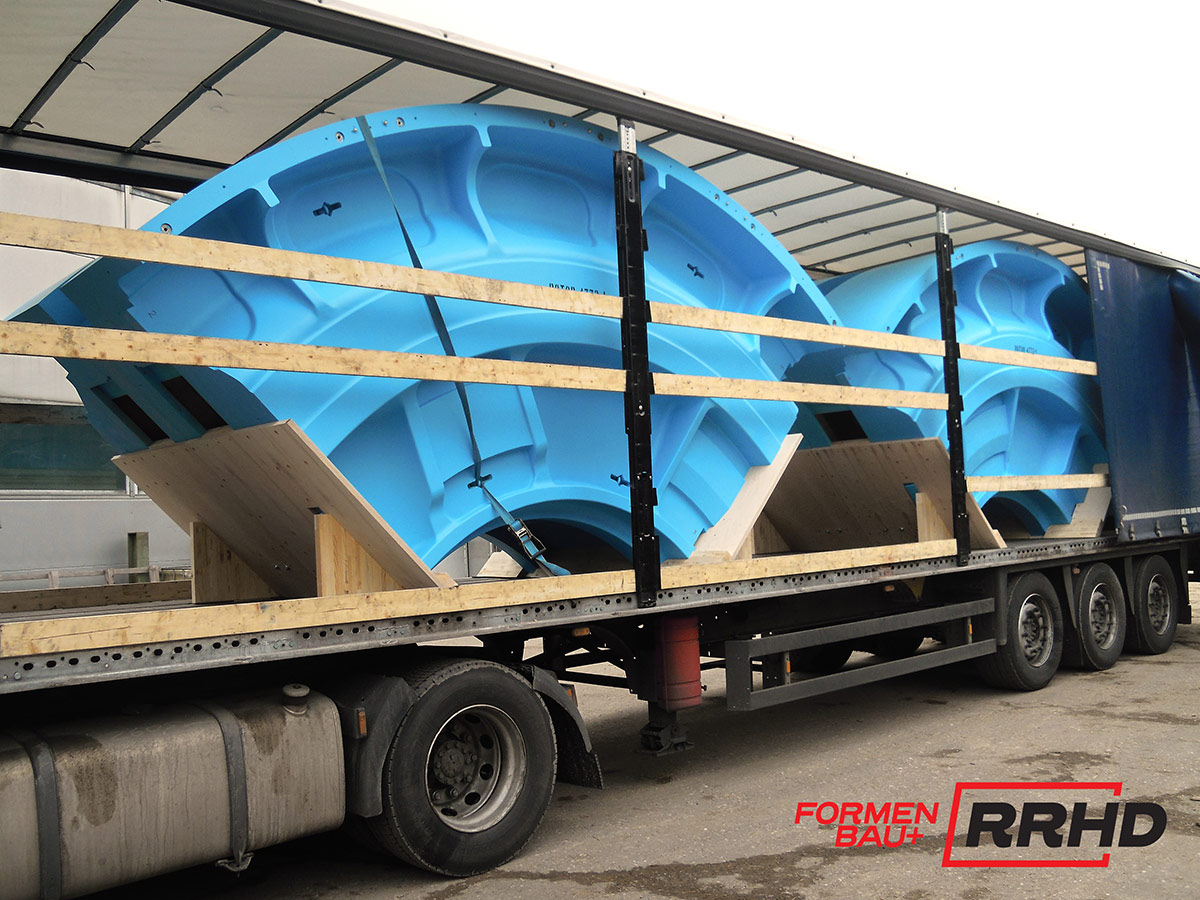 Große, blaue Gießereimodelle mit geschwungener Geometrie und Verstärkungsrippen, sicher verzurrt auf einem Tieflader-Lkw für den Transport. Die Modelle sind auf Holzkonstruktionen gelagert und durch Holzlatten gegen Verrutschen gesichert. Der offene Auflieger gibt den Blick auf mehrere identische Modellsegmente frei.
