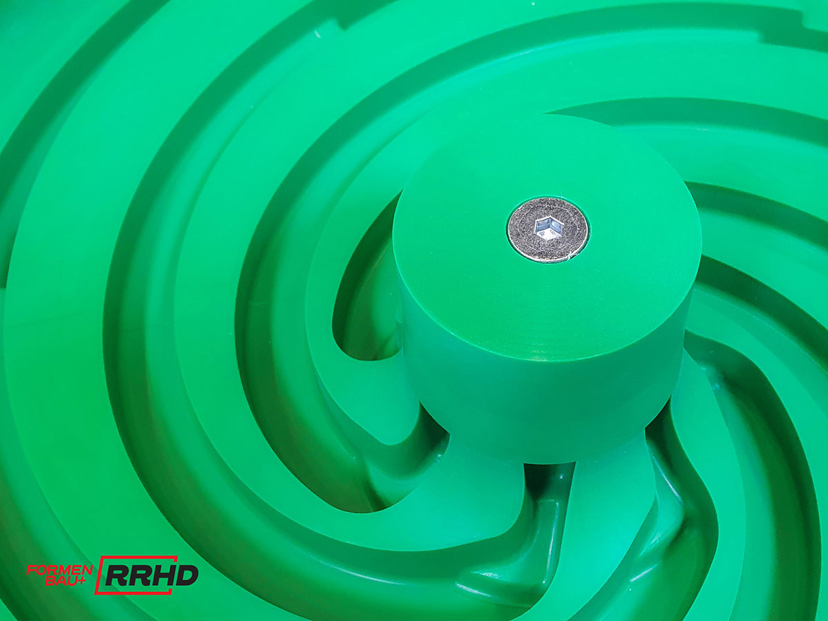 Grünes Gießereimodell mit komplexer, spiralförmiger Struktur und zentralem zylindrischem Element, das mit einer Metallaufnahme versehen ist. Die Geometrie deutet auf ein Laufrad oder eine Pumpenkomponente hin, das Modell wurde präzise bearbeitet und dient als Vorlage für ein rotationssymmetrisches Gussbauteil.