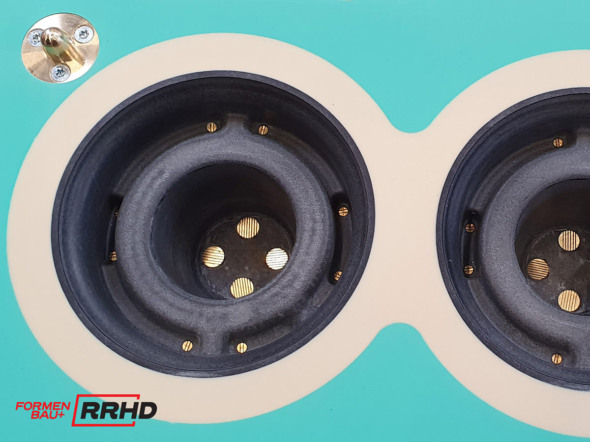 Detailaufnahme eines Gießereimodells mit zwei schwarzen, zylindrisch geformten Hohlräumen in einer türkisfarbenen Trägerplatte. Die Kavitäten sind präzise bearbeitet und mit mehreren Messingdüsen versehen.